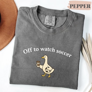 May include: A gray Comfort Colors t-shirt with the text "Off to watch soccer" and a cartoon duck holding a coffee cup. The shirt is a crew neck and has a small tag on the neckline.