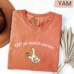 May include: Coral-colored t-shirt with the text "Off to watch soccer." A cartoon duck holding a coffee cup and soccer ball is printed below the text. The shirt is folded and has a Comfort Colors tag.