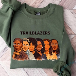 May include: Olive green sweatshirt with the word "TRAILBLAZERS" printed across the top. Below the text is a graphic of six women with varying skin tones. The sweatshirt has long sleeves and a crew neck.