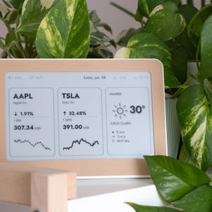 Może przedstawiać: Prostokątny cyfrowy wyświetlacz typu smart z jasnobrązową ramką, prezentujący dane giełdowe dla AAPL i TSLA oraz informacje pogodowe dla Madrytu, w tym temperaturę, warunki nieba i odczyty ciśnienia atmosferycznego na ekranie.