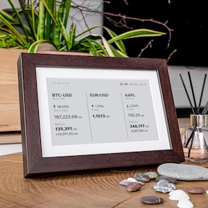 Stock, crypto, forex, ETF & precious metals e-ink live price ticker Brisa Frame rests on a table, encased in a handcrafted wooden frame with soft. The e-ink display shows MSCI ACWI live price (including wallet value), EURUSD currency exchange rate, AAPL stocks price & weather. Designed for slow living and mindful interiors, Brisa blends technology with calm aesthetics, delivering daily information in a serene, non-intrusive way.