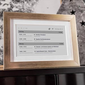 May include: A framed digital display with a gold-colored frame. The screen shows a schedule with text in German, including times and event details. The display is on a dark surface.