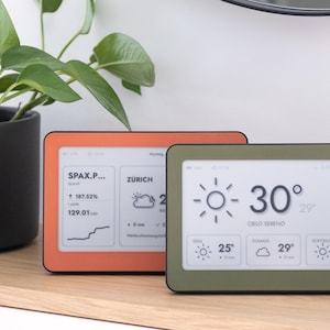 May include: Two rectangular digital weather display devices with screens showing temperature, weather icons, and city names. One device has an orange frame, and the other has an olive green frame. The screens display data including 30 degrees, Roma, and Zurich.