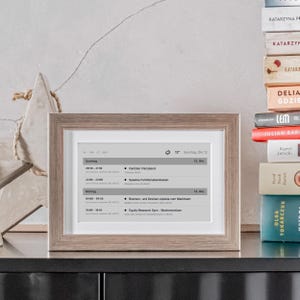 Könnte beinhalten: Ein gerahmter digitaler Kalender zeigt einen Zeitplan mit deutschem Text. Der Rahmen ist hellbraun, und der Bildschirm zeigt eine Liste von Ereignissen für Sonntag und Montag. Ein Stapel Bücher ist im Hintergrund zu sehen.