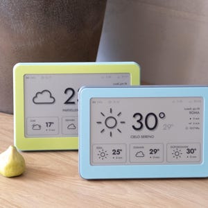 May include: Two rectangular digital weather stations with screens displaying temperature, weather icons, and text. One device has a light green frame, and the other has a light blue frame. The screens show data including 30 degrees and Cielo Sereno.