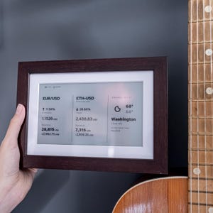 Stock, crypto, forex, ETF & precious metals e-ink live price ticker Brisa Frame rests on a table, encased in a handcrafted wooden frame with soft. The e-ink display shows MSCI ACWI live price (including wallet value), EURUSD currency exchange rate, AAPL stocks price & weather. Designed for slow living and mindful interiors, Brisa blends technology with calm aesthetics, delivering daily information in a serene, non-intrusive way.