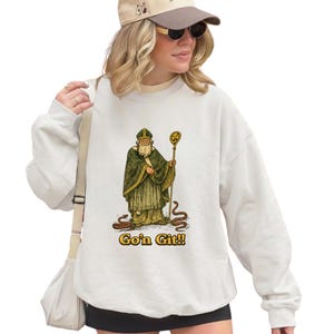 Puede incluir: Sudadera blanca con un gráfico de una figura vestida con una túnica que sostiene un bastón y el texto "Go'n Git!!". La ilustración es en verde, dorado y marrón. La sudadera es de estilo casual.
