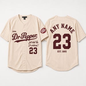 Op de afbeelding: Een beige honkbalshirt met dunne verticale krijtstrepen, voorzien van het bordeauxrode Dr Pepper-logo, de tekst Just what the Dr ordered! en het nummer 23 op de voorzijde. De achterzijde toont de tekst ANY NAME, het nummer 23 en EST. 1885.