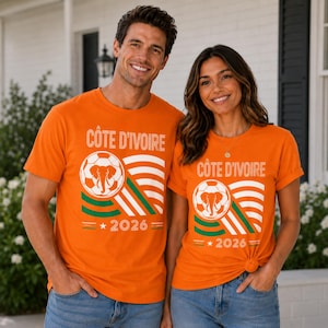 May include: An orange short-sleeve t-shirt featuring the text CÔTE D'IVOIRE, a graphic of a soccer ball with an elephant silhouette inside, curved stripes in green, white, and orange, and the year 2026 with a star on the front.