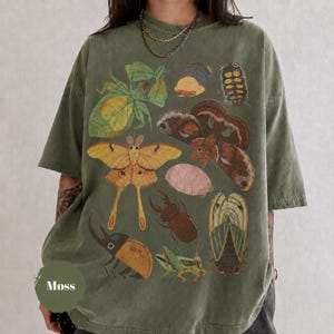 Peut inclure: Un t-shirt à manches courtes vert olive avec une impression graphique représentant divers insectes et papillons dans des tons de brun, jaune et vert. Le motif comprend un grand papillon jaune, un scarabée brun et d'autres illustrations d'insectes.