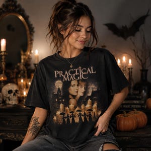 Könnte beinhalten: Ein schwarzes Kurzarm-T-Shirt mit einem Grafikdruck und dem Text Practical Magic, das zwei Frauen, ein großes Haus, einen Vollmond und eine Reihe von Silhouetten mit Regenschirmen vor zahlreichen brennenden Kerzen zeigt.