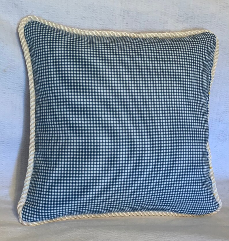 Pu&ograve; includere: Un cuscino in tessuto gingham blu e bianco con un bordo in corda bianca.