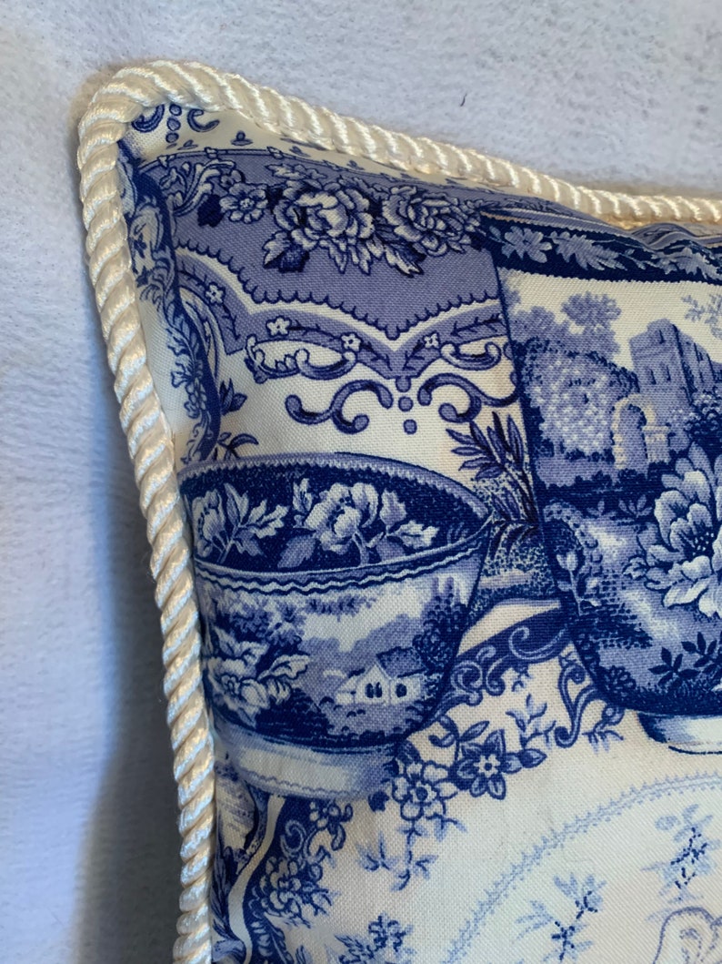 Pu&ograve; includere: Un cuscino bianco con un motivo floreale blu e bianco. Il cuscino ha un bordo in corda bianca attorno ai bordi.