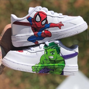 Two custom kids Nike Air Force 1 Low sneakers stacked together featuring hand-painted Marvel characters—Spider-Man in red and blue on the top shoe, Hulk in bright green on the bottom shoe, both with bold black outlines on white leather