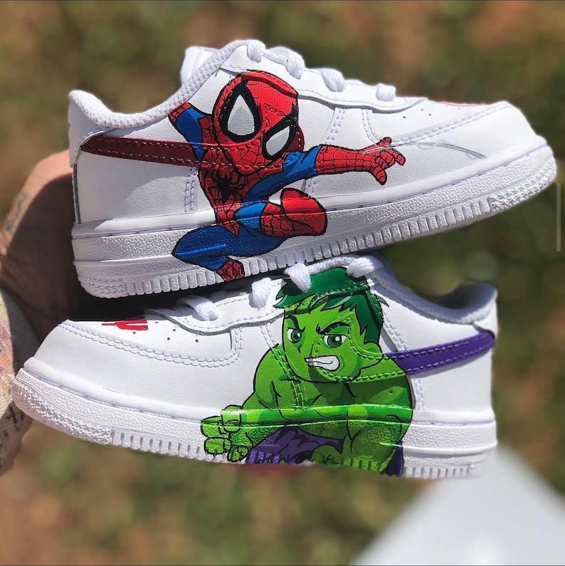 Two custom kids Nike Air Force 1 Low sneakers stacked together featuring hand-painted Marvel characters—Spider-Man in red and blue on the top shoe, Hulk in bright green on the bottom shoe, both with bold black outlines on white leather
