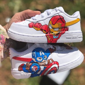 Close-up of custom kids Marvel Nike Air Force 1 shoes held in hand, showing Iron Man in red and gold armor on top shoe and Captain America in blue with red shield on bottom shoe, hand-painted on white leather with detailed character artwo