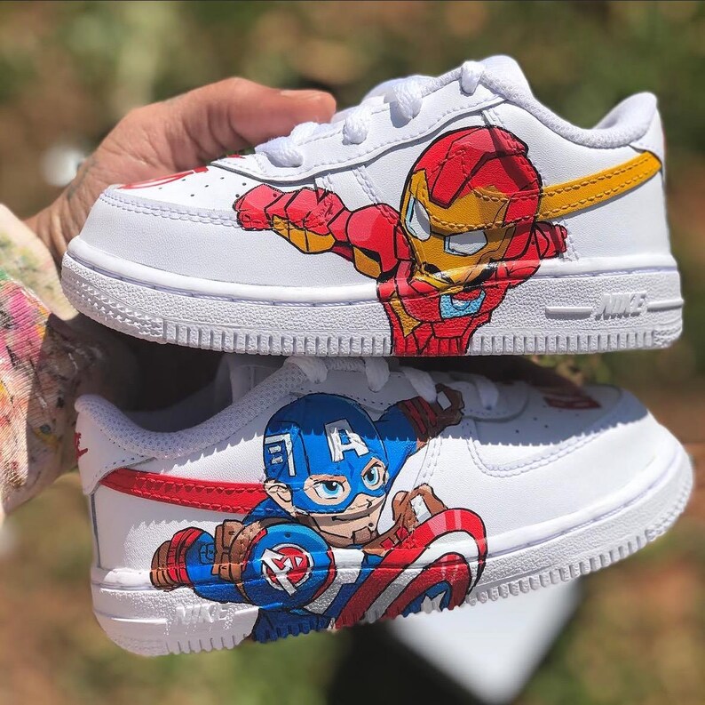 Close-up of custom kids Marvel Nike Air Force 1 shoes held in hand, showing Iron Man in red and gold armor on top shoe and Captain America in blue with red shield on bottom shoe, hand-painted on white leather with detailed character artwo