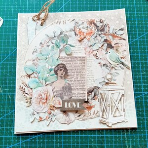 May include: A square greeting card featuring a collage of a vintage woman, a white lantern, a small bird, a butterfly, and floral elements. The design includes a piece of printed text, a banner with the word LOVE, and a jute twine loop at the top.