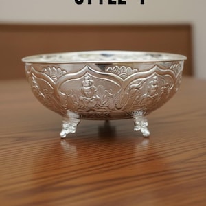 May include: A silver-coloured metal bowl featuring embossed figures and floral patterns on its exterior. The bowl is supported by three small, ornate feet and has the text STYLE 4 printed in black above it. The surface is reflective and metallic.