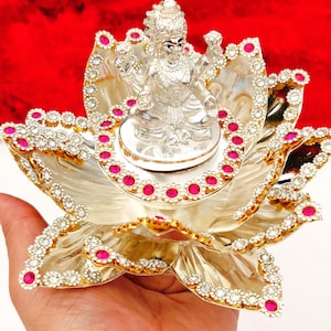 以下が含まれることがあります： 銀色の蓮の花の上に、神様の銀色の像が置かれています。蓮の花びらは、透明とピンクの宝石で縁取られています。背景は無地の赤色です。