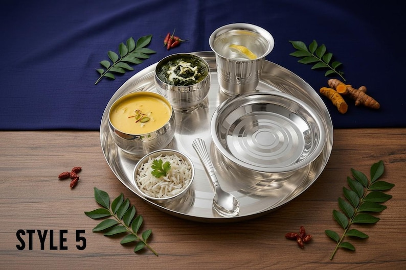 May include: A stainless steel thali set featuring a large round plate, three small bowls, a drinking glass, and a spoon. The set includes a bowl of yellow soup, a bowl of green vegetable curry, and a bowl of white rice. Text reads STYLE 5.