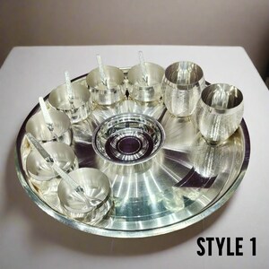 May include: A silver-colored metal dinner set featuring a large round serving tray, a central bowl, four small bowls with matching spoons, and two tumblers. The items have a polished finish with subtle etched line patterns. Text reads STYLE 1.