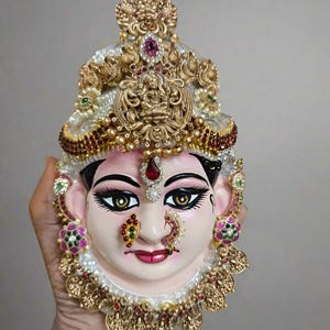 Puede incluir: Una máscara decorativa de una deidad con una expresión serena. La máscara presenta una elaborada ornamentación dorada, que incluye una corona enjoyada, aretes y un anillo en la nariz. La cara está pintada con rasgos delicados, incluidos ojos oscuros y labios rojos. El texto "STYLE 6" está en la parte superior.