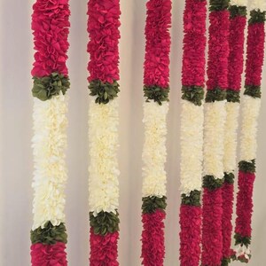 May include: Vertical garlands of cream-colored and magenta flowers hang against a white wall. The garlands are segmented with green and cream-colored accents. The flowers appear to be made of fabric or paper, creating a decorative display.