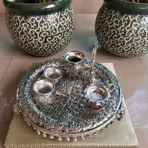 Op de afbeelding: Een zilverkleurige serveerschaal met ingewikkelde details, met kleine kommen en een decoratieve container. De schaal staat op een goudkleurige, beige rechthoekige basis. Twee groen-wit gepatroneerde keramische potten met planten op de achtergrond.