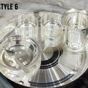 May include: A silver-colored dinnerware set labeled STYLE 6, consisting of a large round tray, two small bowls, one larger bowl with a matching spoon, and a tall drinking glass. Each piece features a polished, reflective metallic finish.