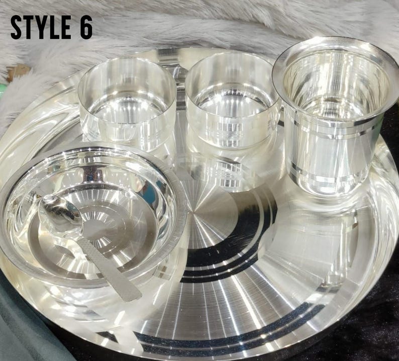 May include: A silver-colored dinnerware set labeled STYLE 6, consisting of a large round tray, two small bowls, one larger bowl with a matching spoon, and a tall drinking glass. Each piece features a polished, reflective metallic finish.