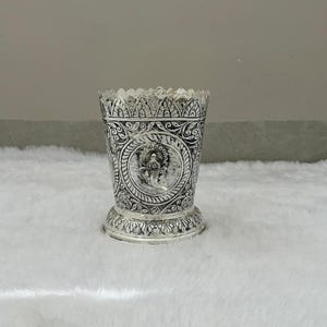 May include: A silver-colored cylindrical vessel featuring an intricate embossed pattern throughout its surface. The center of the vessel displays a circular medallion with a relief of a deity, while the top and bottom edges have scalloped borders.