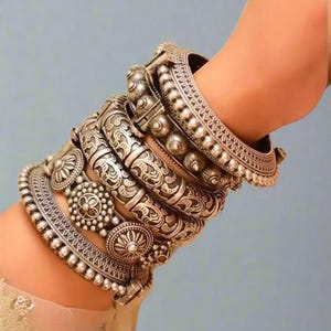 May include: A collection of silver bangles with intricate floral and geometric patterns. The bangles vary in width and design, featuring beaded edges and embossed details. The jewelry is stacked on a wrist, showcasing the ornate craftsmanship.