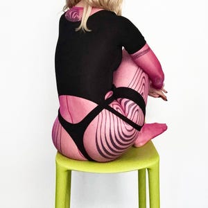Puede incluir: Una persona con un body negro con medias y mangas rosas estampadas, sentada en un taburete verde lima. Las medias y las mangas tienen un diseño ondulado y a rayas. La persona tiene el pelo rubio y un tatuaje en la espalda.
