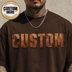 May include: A brown t-shirt with the word "CUSTOM" in a distressed, rusty-orange font across the chest. A circular patch with the text "CUSTOM HERE" and "PREMIUM QUALITY" is on the upper left. A silver chain necklace is visible.