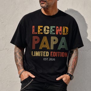 May include: Black t-shirt with the text "LEGEND PAPA LIMITED EDITION EST. 2026" in colorful, distressed lettering. The shirt has a classic crew neck and short sleeves. The design features a vintage, retro style.