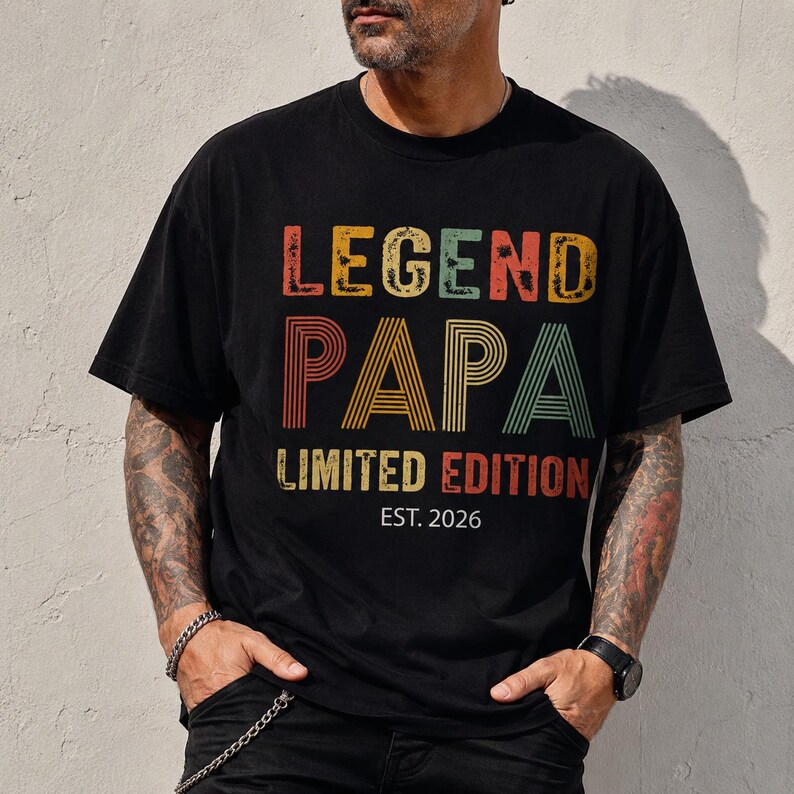 May include: Black t-shirt with the text "LEGEND PAPA LIMITED EDITION EST. 2026" in colorful, distressed lettering. The shirt has a classic crew neck and short sleeves. The design features a vintage, retro style.