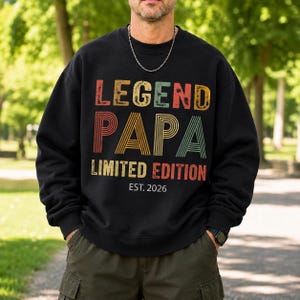 May include: Black sweatshirt with the text "LEGEND PAPA LIMITED EDITION EST. 2026" in a retro color scheme. The text is in a bold, blocky font. The sweatshirt is worn by a person in an outdoor setting.