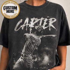 May include: Dark grey t-shirt with a baseball player graphic swinging a bat. The name "CARTER" is printed in white above the player. The shirt also has a "Custom Design" patch.
