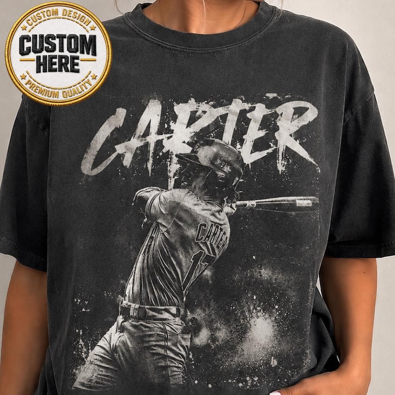 May include: Dark grey t-shirt with a baseball player graphic swinging a bat. The name "CARTER" is printed in white above the player. The shirt also has a "Custom Design" patch.