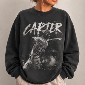May include: Charcoal grey sweatshirt with a vintage-style baseball graphic. The design features a baseball player swinging a bat, with the name "CARTER" in a distressed white font. A great gift for sports enthusiasts.
