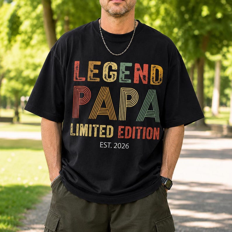 May include: Black t-shirt with the text "LEGEND PAPA LIMITED EDITION EST. 2026" in a retro font. The shirt has a crew neck and short sleeves. The person is wearing a silver chain and a watch.
