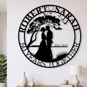 May include: Black metal wall art featuring a couple kissing under a tree, with the names "Robert" and "Sarah" and the text "50 Years Together" and "Est. 1974" in a circular design. The art is mounted on a white wall above a wooden console.