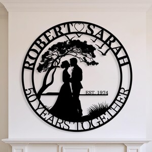Può includere: Una decorazione da parete circolare in metallo nero con la silhouette di una coppia sotto un albero. Il testo riporta ROBERT, SARAH, 50 YEARS TOGETHER e EST. 1974. Una piccola icona a forma di cuore è posizionata al centro della cornice.