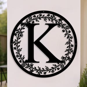 May include: Black metal wall art featuring the letter "K" inside a circular frame. The letter is centered, and the frame is adorned with a floral wreath design. The artwork is mounted on a white wall, suitable for indoor or outdoor decor.