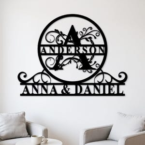 Peut inclure: Décoration murale en métal noir avec la lettre "A" dans un cercle, le nom de famille "ANDERSON" et les noms "ANNA & DANIEL" en dessous. Le design comprend des volutes décoratives et est monté sur un mur blanc.