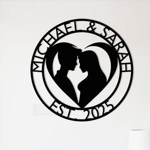 May include: Black metal wall art depicting a couple's silhouettes inside a heart shape. The names "MICHAEL & SARAH" are inscribed around the heart, with "EST. 2025" at the bottom. The art is mounted on a white wall.