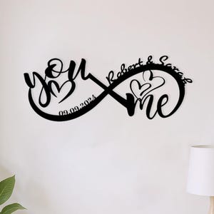 Può includere: Una decorazione da parete in metallo nero a forma di simbolo dell'infinito con le parole you e me, i nomi Robert & Sarah e la data 09.09.2024. Il design include due contorni di cuori integrati nella scritta in corsivo e nell'anello dell'infinito.