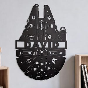 Peut inclure: Une décoration murale en métal noir en forme de vaisseau spatial Millennium Falcon, présentant des détails mécaniques découpés complexes et le nom DAVID affiché en évidence dans une découpe rectangulaire horizontale au centre.