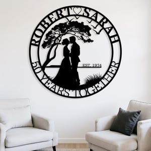 May include: A circular black metal wall sign featuring the names ROBERT and SARAH at the top with a heart icon. The center shows a silhouette of a couple under a tree, with the text EST. 1974 and 50 YEARS TOGETHER along the bottom edge.